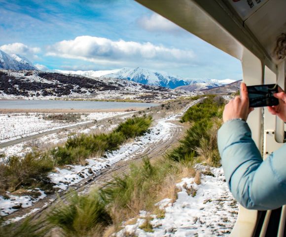 View from a train window of a snowy landscape with mountains in the distance. Person in blue coat takes photo, capturing serene, scenic beauty.