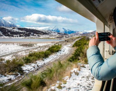 View from a train window of a snowy landscape with mountains in the distance. Person in blue coat takes photo, capturing serene, scenic beauty.