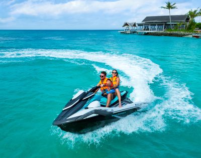 A couple rides a jet ski in clear turquoise waters near a tropical resort. They wear orange life vests, smiling under a vibrant blue sky.