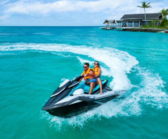 A couple rides a jet ski in clear turquoise waters near a tropical resort. They wear orange life vests, smiling under a vibrant blue sky.