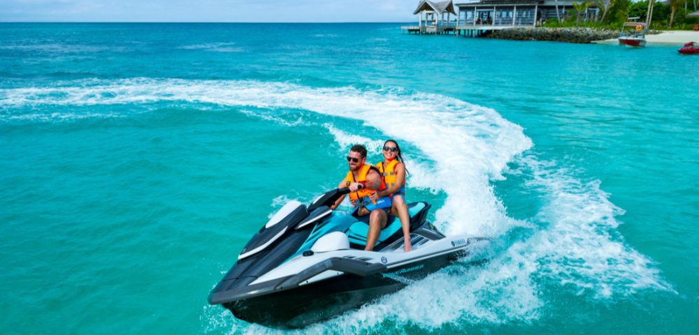 A couple rides a jet ski in clear turquoise waters near a tropical resort. They wear orange life vests, smiling under a vibrant blue sky.
