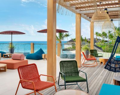 Outdoor patio overlooking the ocean with colorful chairs, bean bags, and umbrellas. Wooden pergola above creates a relaxed, tropical vibe.
