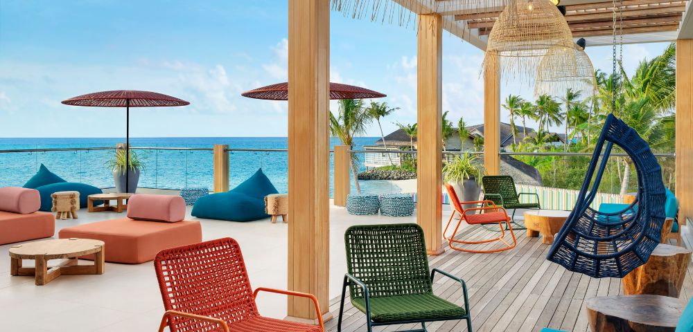 Outdoor patio overlooking the ocean with colorful chairs, bean bags, and umbrellas. Wooden pergola above creates a relaxed, tropical vibe.