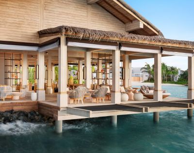 A luxurious overwater bungalow with open wooden design, surrounded by turquoise ocean. The elegant interior features cozy seating and natural decor.