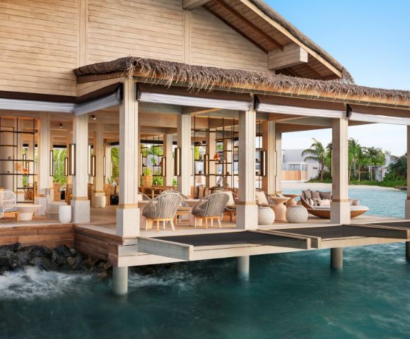 A luxurious overwater bungalow with open wooden design, surrounded by turquoise ocean. The elegant interior features cozy seating and natural decor.