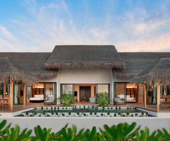Luxurious beachfront villa with palm thatched roof, flanked by open verandas. A pristine pool reflects vibrant greenery under a clear blue sky, creating a tranquil atmosphere.