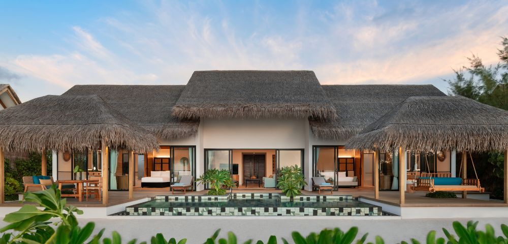 Luxurious beachfront villa with palm thatched roof, flanked by open verandas. A pristine pool reflects vibrant greenery under a clear blue sky, creating a tranquil atmosphere.