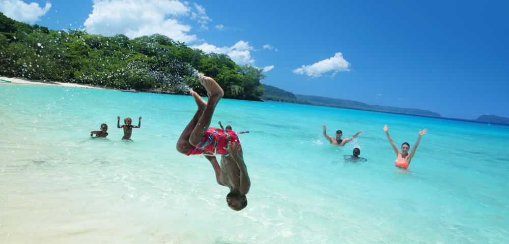 A group of people joyfully jumping into the ocean on a sunny day, splashing water around them.