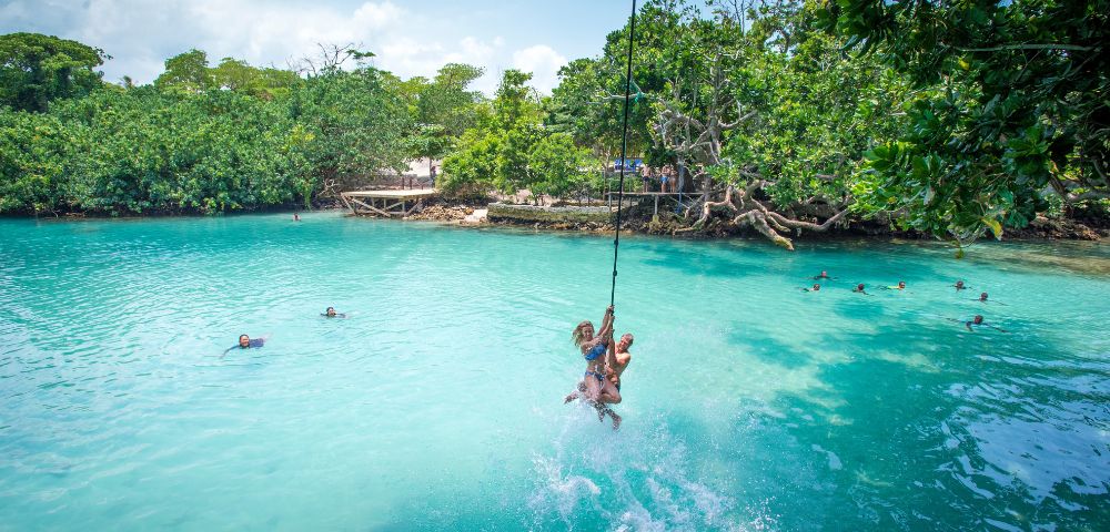 People swing on a rope over clear blue water, splashing down while others swim nearby; lush green trees surround the area under a partly cloudy sky.