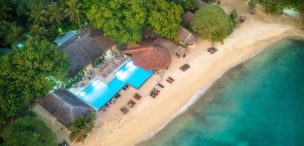 Aerial view of a beachfront resort with a large pool, sun loungers, thatched-roof buildings, sandy beach, and turquoise water surrounded by dense green trees.