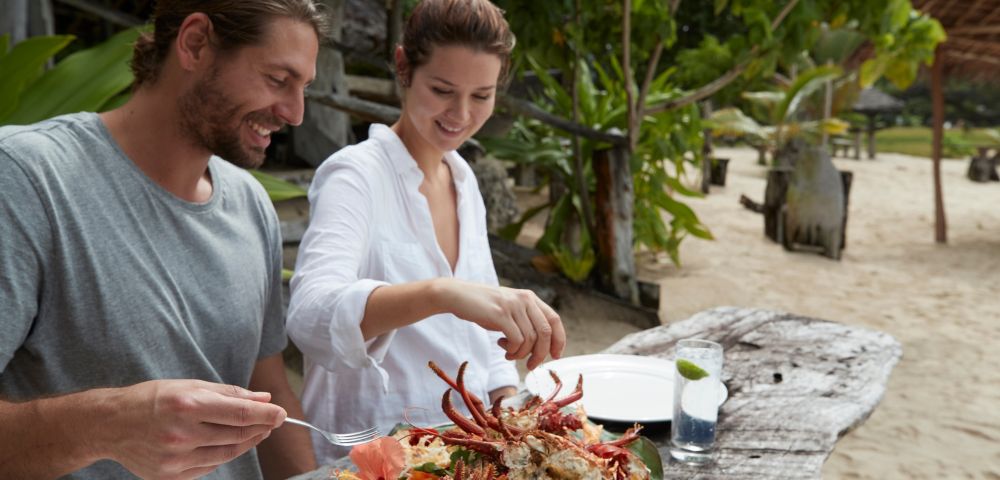 A man and woman enjoying a meal together at a table on the beach, with waves gently lapping in the background.