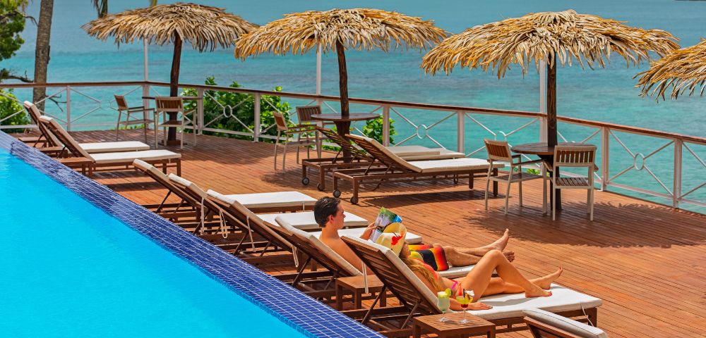 A man and woman relaxing on a lounge chair by a pool with the ocean in the background.