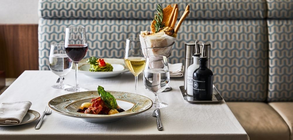 vA restaurant table with elegant plating features a pasta dish, red and white wine, fresh salad, and breadsticks, set on a white tablecloth, creating a refined ambiance.