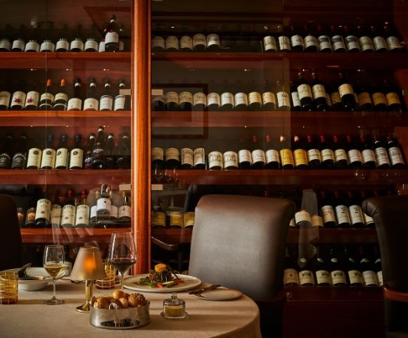 Cozy restaurant interior with a wall of wine bottles behind a set dining table. A warm ambiance is created by dim lighting and elegant tableware.