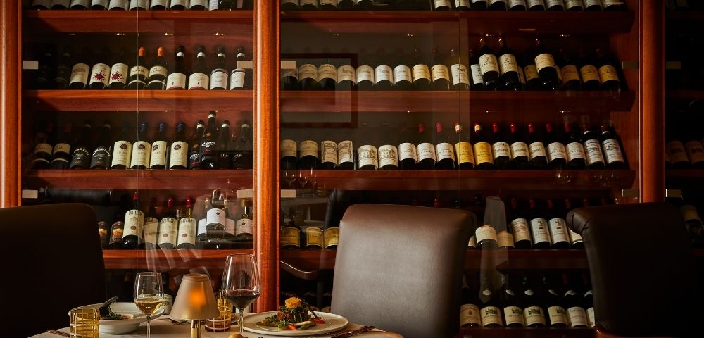 Cozy restaurant interior with a wall of wine bottles behind a set dining table. A warm ambiance is created by dim lighting and elegant tableware.