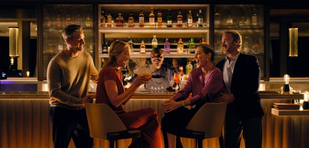 A group of four people dressed elegantly enjoy a lively conversation at a dimly lit bar, with shelves of bottles glowing warmly in the background.