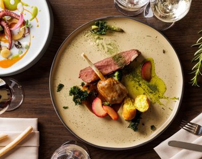 A gourmet dish featuring lamb chops, sliced steak, roasted potatoes, and greens on a beige plate, with a side of seafood salad, exuding elegance.