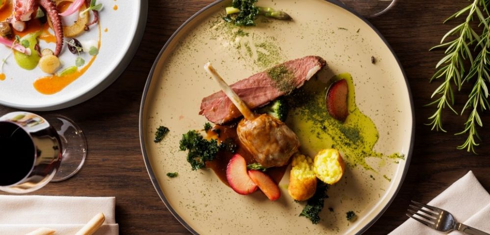 A gourmet dish featuring lamb chops, sliced steak, roasted potatoes, and greens on a beige plate, with a side of seafood salad, exuding elegance.