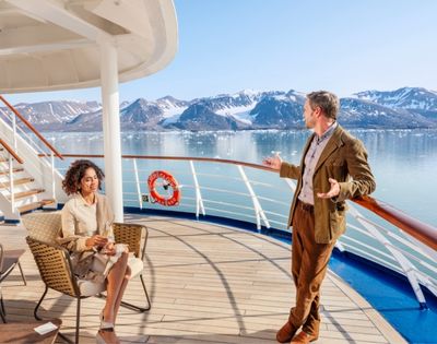 A man and woman enjoy a sunny day on a ship's deck, surrounded by icy mountains and calm water. The scene conveys relaxation and awe at nature's beauty.