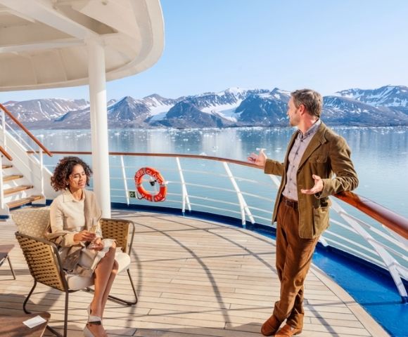 A man and woman enjoy a sunny day on a ship's deck, surrounded by icy mountains and calm water. The scene conveys relaxation and awe at nature's beauty.