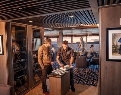 A group of people are inside a luxurious cabin with a modern design. Two men examine documents by a cabinet, while others relax, enjoying an ocean view at sunset.
