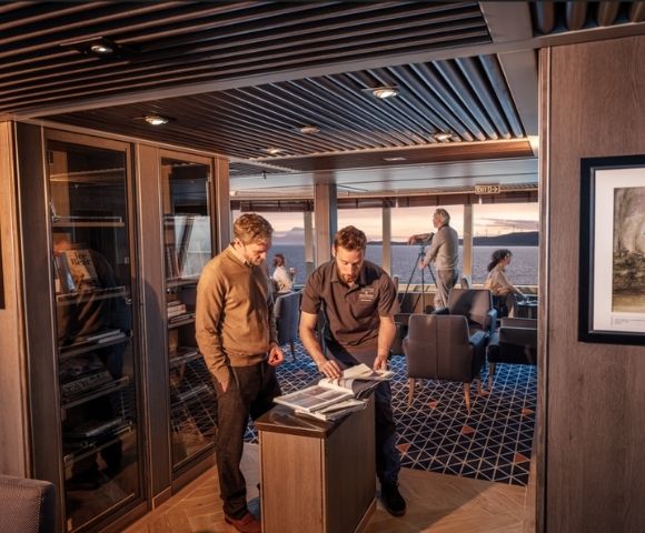 A group of people are inside a luxurious cabin with a modern design. Two men examine documents by a cabinet, while others relax, enjoying an ocean view at sunset.