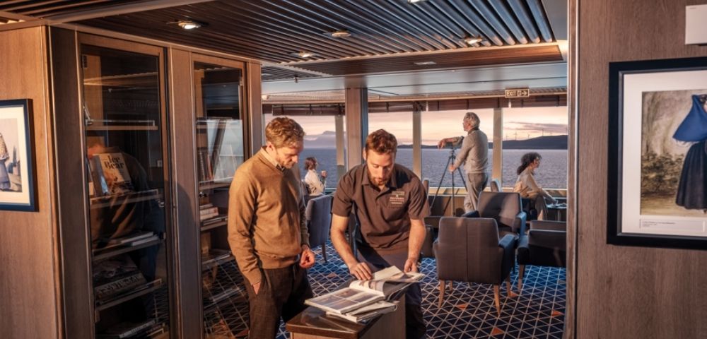 A group of people are inside a luxurious cabin with a modern design. Two men examine documents by a cabinet, while others relax, enjoying an ocean view at sunset.