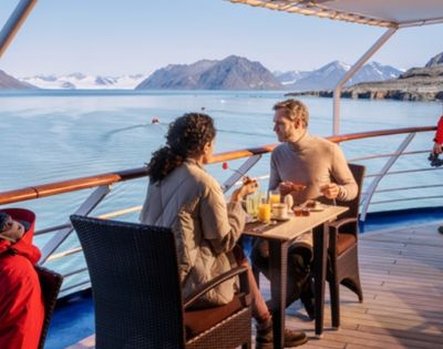 A couple enjoys breakfast on a ship's deck, overlooking calm waters and distant snow-capped mountains under a clear blue sky, creating a serene atmosphere.