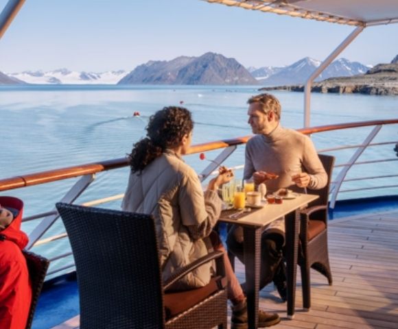 A couple enjoys breakfast on a ship's deck, overlooking calm waters and distant snow-capped mountains under a clear blue sky, creating a serene atmosphere.