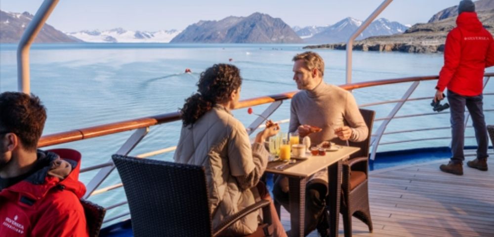 A couple enjoys breakfast on a ship's deck, overlooking calm waters and distant snow-capped mountains under a clear blue sky, creating a serene atmosphere.