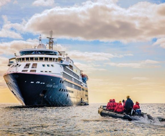 A large cruise ship on the ocean approaches the horizon under a cloudy sky. In the foreground, a small boat with people in red jackets sails nearby.