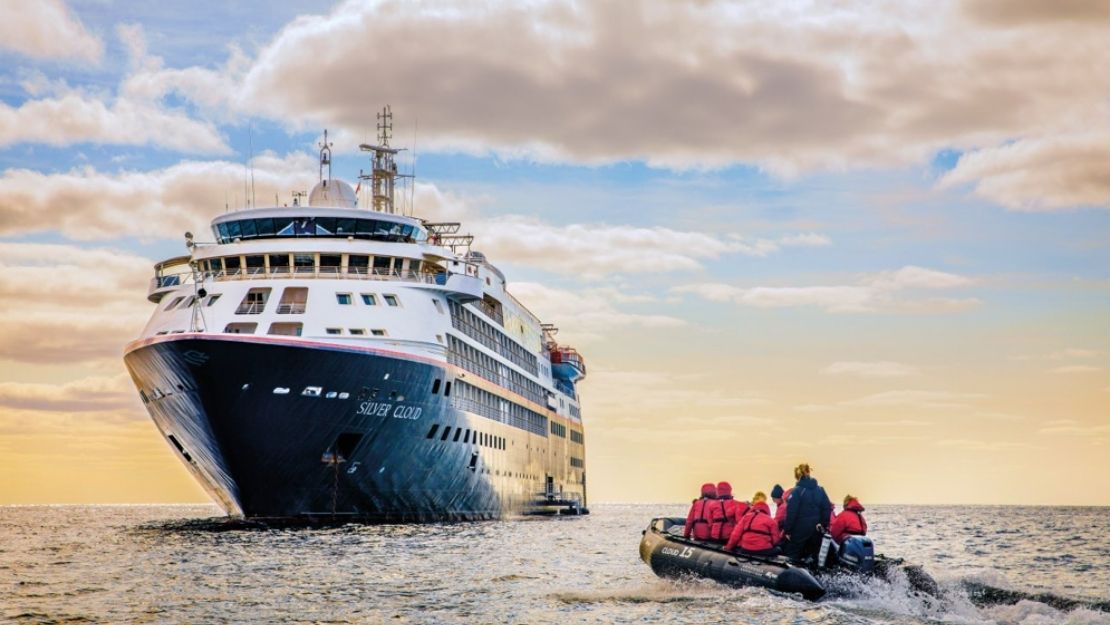 A large cruise ship on the ocean approaches the horizon under a cloudy sky. In the foreground, a small boat with people in red jackets sails nearby.