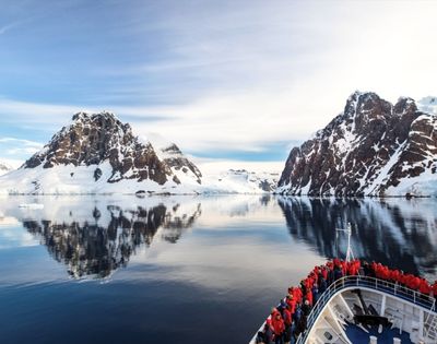 A ship with passengers in red jackets approaches calm, reflective waters surrounded by snow-covered mountains under a clear, blue sky.