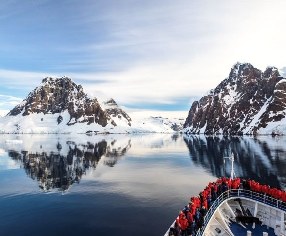 A ship with passengers in red jackets approaches calm, reflective waters surrounded by snow-covered mountains under a clear, blue sky.