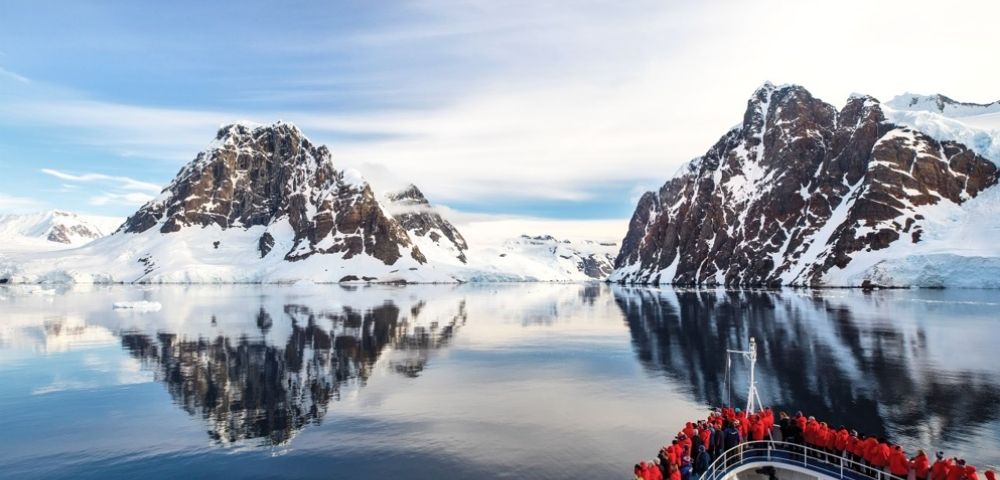 A ship with passengers in red jackets approaches calm, reflective waters surrounded by snow-covered mountains under a clear, blue sky.