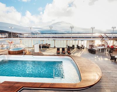 Sunny cruise ship deck featuring a pool and lounge chairs, with a hot tub nearby. Snow-capped mountains and blue sky create a serene backdrop.