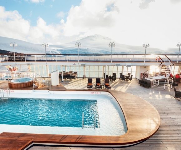 Sunny cruise ship deck featuring a pool and lounge chairs, with a hot tub nearby. Snow-capped mountains and blue sky create a serene backdrop.