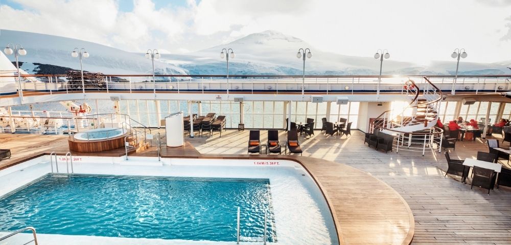 Sunny cruise ship deck featuring a pool and lounge chairs, with a hot tub nearby. Snow-capped mountains and blue sky create a serene backdrop.