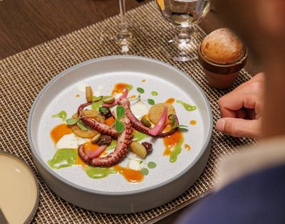 Elegant dish with octopus tentacles, pickled vegetables, and colorful sauces on a white plate. Served on a woven mat with a small bread roll and wine glass nearby.