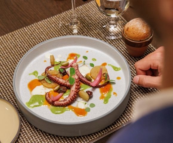 Elegant dish with octopus tentacles, pickled vegetables, and colorful sauces on a white plate. Served on a woven mat with a small bread roll and wine glass nearby.