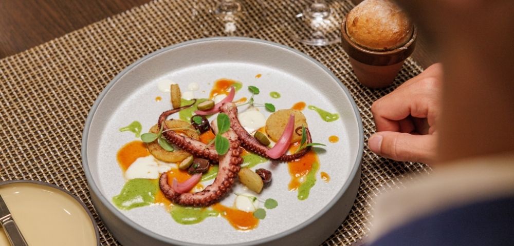 Elegant dish with octopus tentacles, pickled vegetables, and colorful sauces on a white plate. Served on a woven mat with a small bread roll and wine glass nearby.