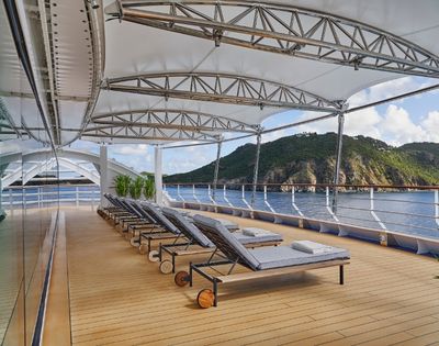 Luxurious yacht deck with six gray chaise lounges under a canopy. Calm sea and lush green hills in the background, exuding relaxation and tranquility.