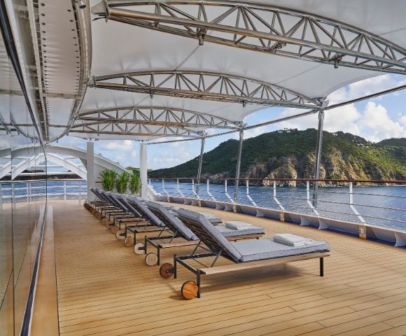 Luxurious yacht deck with six gray chaise lounges under a canopy. Calm sea and lush green hills in the background, exuding relaxation and tranquility.