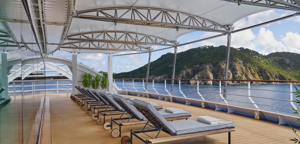 Luxurious yacht deck with six gray chaise lounges under a canopy. Calm sea and lush green hills in the background, exuding relaxation and tranquility.