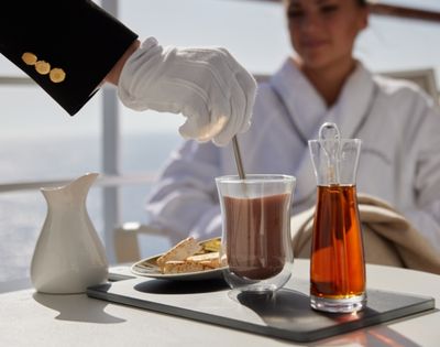 A person in white gloves stirs a beverage on a tray, with a jug, glass, and carafe. A woman in a robe watches, creating a serene, luxurious atmosphere.