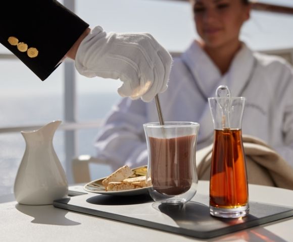 A person in white gloves stirs a beverage on a tray, with a jug, glass, and carafe. A woman in a robe watches, creating a serene, luxurious atmosphere.