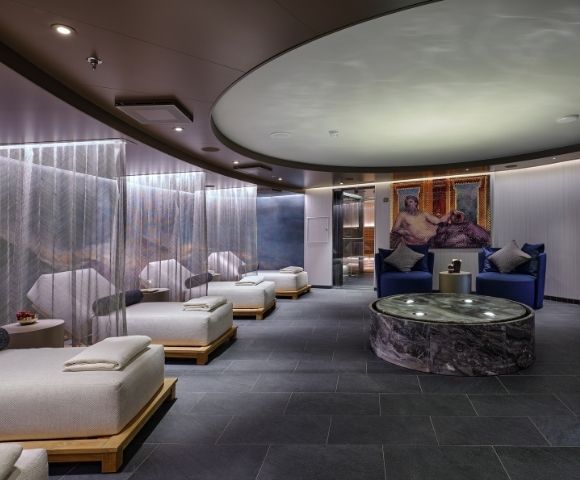 Luxurious spa lounge with a serene ambiance. Features beige cushioned loungers, soft lighting, circular ceiling design, and a marble table. Calm and inviting.