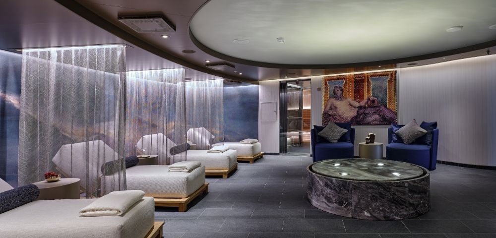 Luxurious spa lounge with a serene ambiance. Features beige cushioned loungers, soft lighting, circular ceiling design, and a marble table. Calm and inviting.
