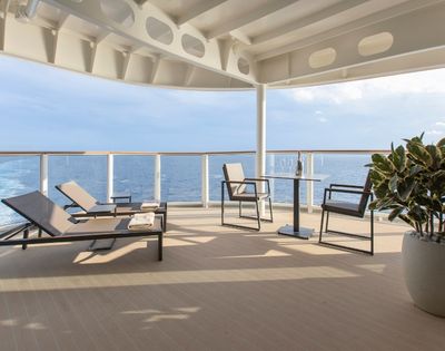 Spacious cruise ship deck with modern loungers and chairs, a small table, and a potted plant. Ocean views and clear blue sky create a serene atmosphere.