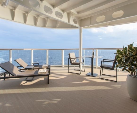 Spacious cruise ship deck with modern loungers and chairs, a small table, and a potted plant. Ocean views and clear blue sky create a serene atmosphere.